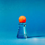 A small clear blue glass bottle or vial with a rounded orange cap, photographed on a smooth blue studio background.