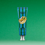 A blue and green striped tube of SOFTY HAND CREAM PISTACHIO stands upright on a green background, featuring a pistachio illustration on the front. This serum-in-cream hand moisturizer delivers hydration with a silky finish.