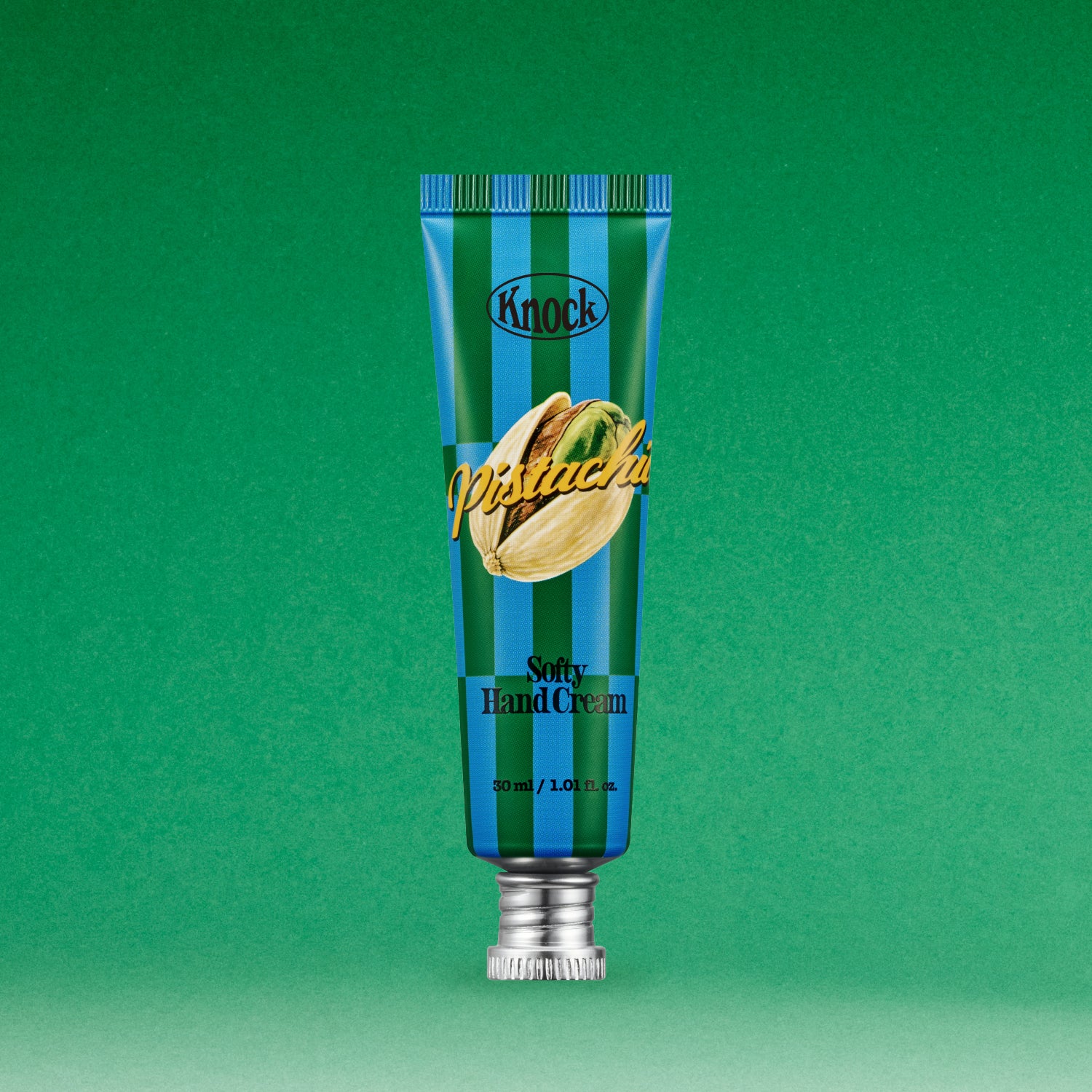 A blue and green striped tube of SOFTY HAND CREAM PISTACHIO stands upright on a green background, featuring a pistachio illustration on the front. This serum-in-cream hand moisturizer delivers hydration with a silky finish.