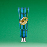 A blue and green striped tube of SOFTY HAND CREAM PISTACHIO stands upright on a green background, featuring a pistachio illustration on the front. This serum-in-cream hand moisturizer delivers hydration with a silky finish.