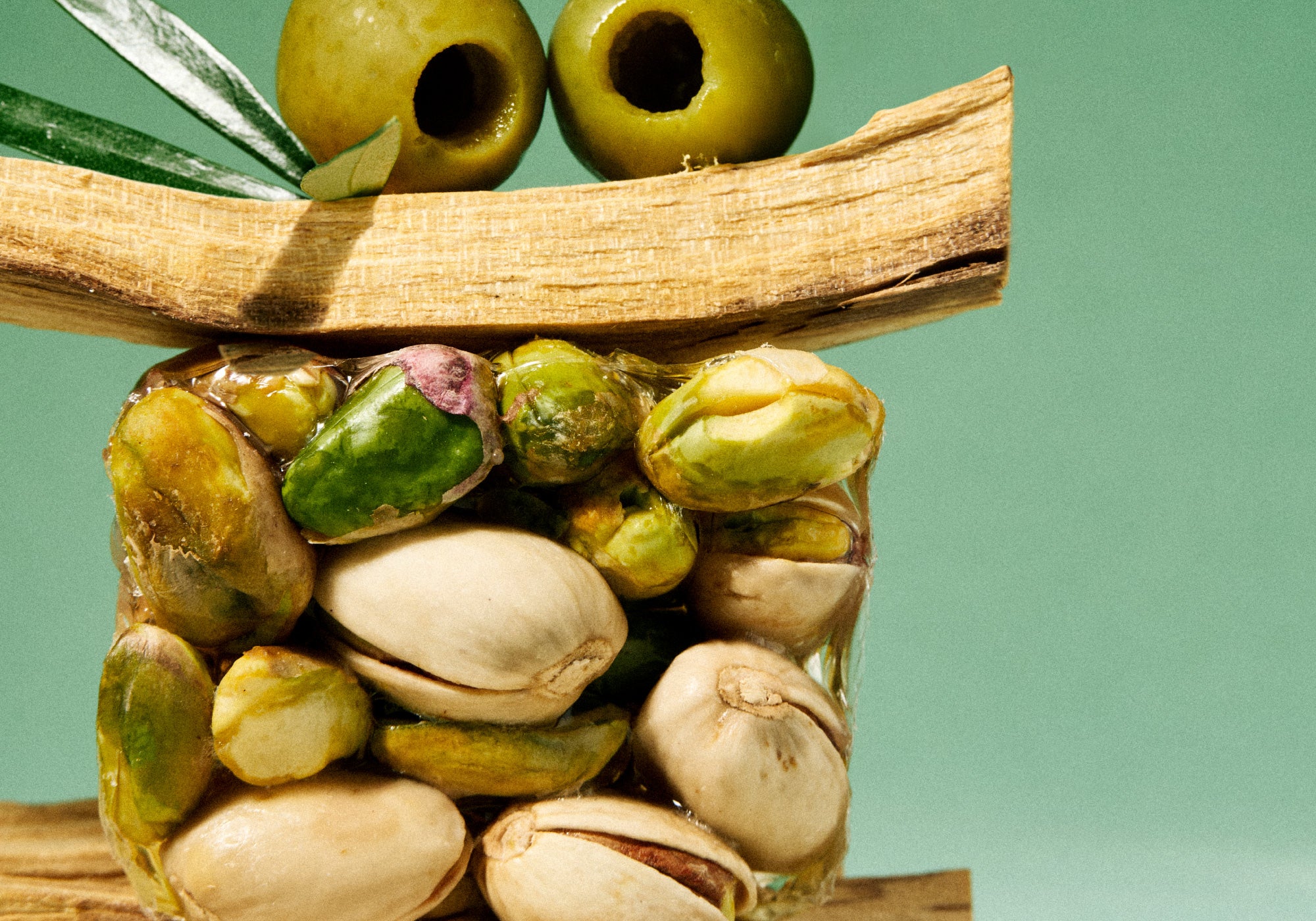 A stack of pistachios and green olives is artistically arranged to resemble a face with olives as eyes against a green background.