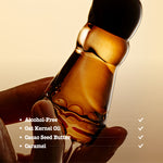 A hand holds a small clear bottle filled with amber-colored oil against a warm beige background. White text on the left lists product attributes: Alcohol-Free, Oat Kernel Oil, Cacao Seed Butter, and Caramel, with checkmarks on the right.