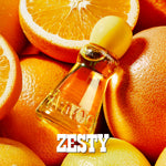 A small clear glass perfume bottle filled with amber-orange liquid, topped with a pale yellow spherical cap, surrounded by sliced oranges and whole citrus fruits. The word 'ZESTY' appears in large white letters at the bottom.