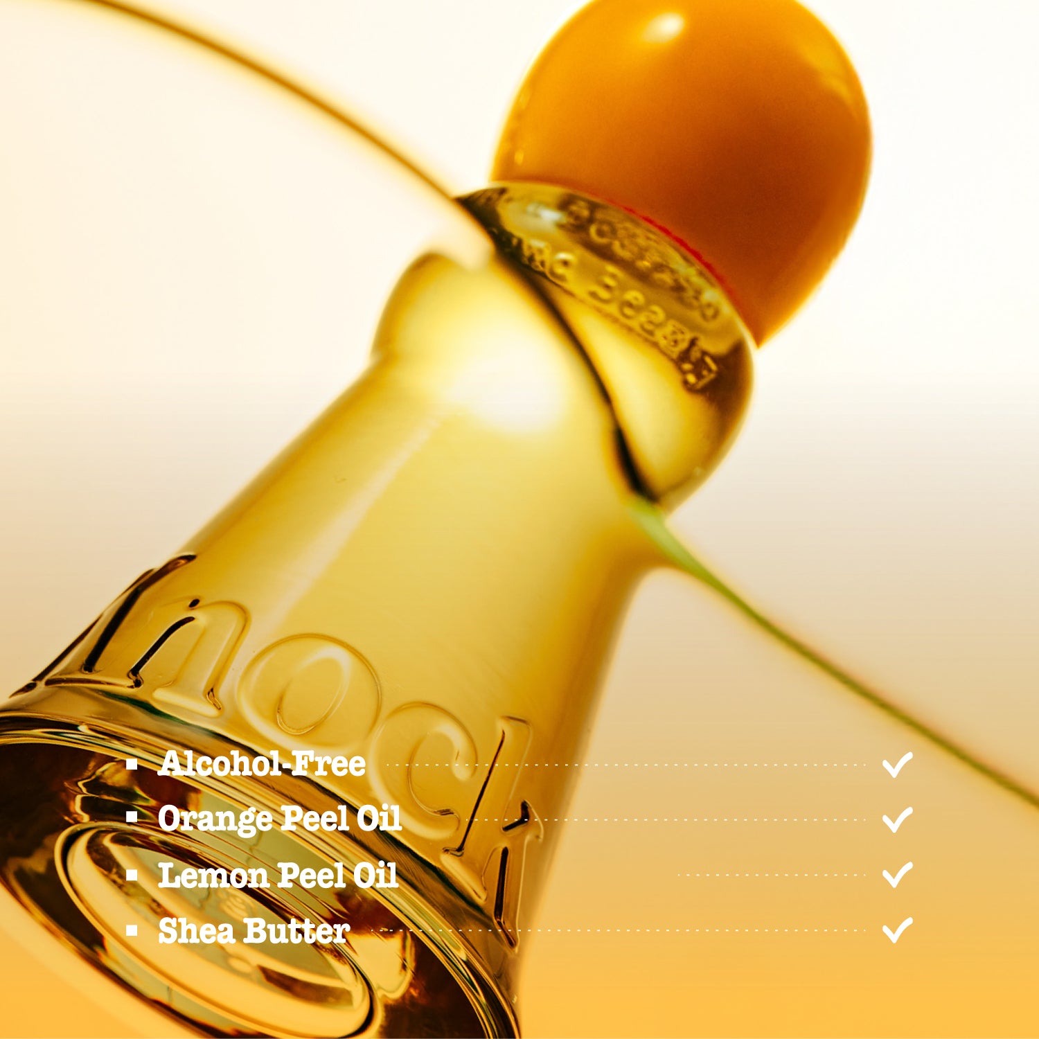 A close-up of a small cosmetic bottle filled with golden yellow oil, with a bright orange cap. White text on the image lists ingredients: Alcohol Free, Orange Peel Oil, Lemon Peel Oil, and Shea Butter.