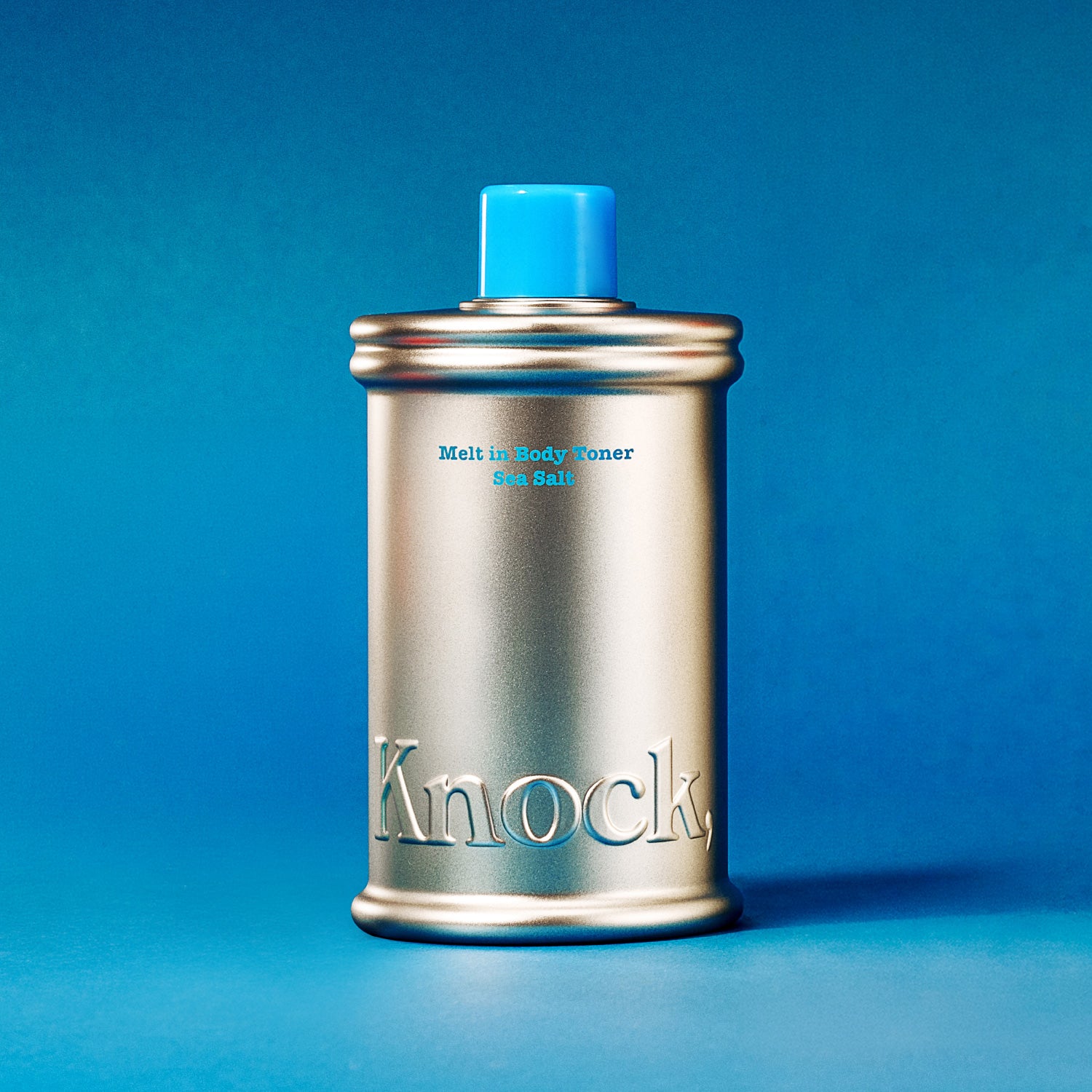 A silver canister with a blue cap, embossed with “Knock” at the bottom and labeled “MELT IN BODY TONER SEA SALT,” is displayed against a blue gradient background. This moisturizing MELT IN BODY TONER absorbs instantly, leaving your skin refreshed and pleasantly scented for hours.