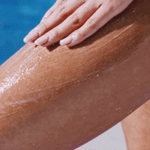 A hand applies lotion or sunscreen to a person’s thigh, with a pool of blue water in the background, leaving the skin looking smooth—perfectly prepped after using SANDY SCRUB WASH.