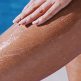 A hand applies lotion or sunscreen to a person’s thigh, with a pool of blue water in the background, leaving the skin looking smooth—perfectly prepped after using SANDY SCRUB WASH.