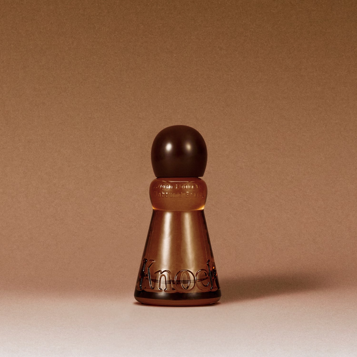 A frosted amber glass perfume bottle with a dark brown spherical cap, featuring the brand name 'Knock' embossed on the front, set against a solid warm brown background.