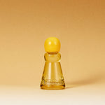 A small translucent amber glass bottle with a rounded yellow cap, centered on a warm beige gradient background. The bottle has embossed lettering near the base.
