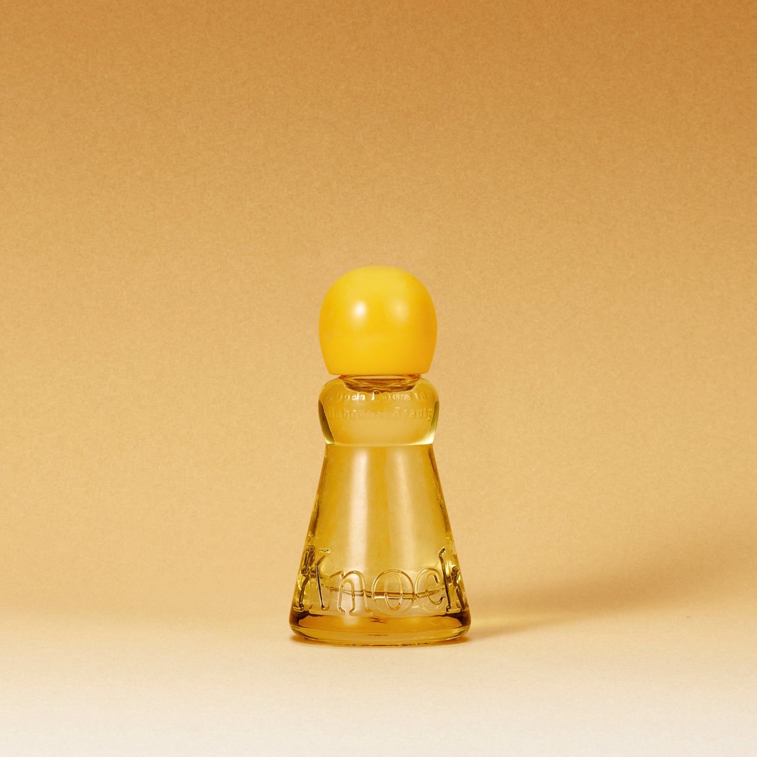 A small translucent amber glass bottle with a rounded yellow cap, centered on a warm beige gradient background. The bottle has embossed lettering near the base.