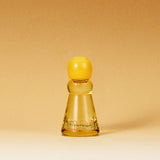 A small translucent amber glass bottle with a rounded yellow cap, centered on a warm beige gradient background. The bottle has embossed lettering near the base.