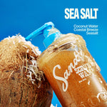 A bottle of SANDY SCRUB WASH with a blue pump sits next to a cracked coconut, coconut shavings scattered nearby. Text highlights SEA SALT, Coconut Water, Coastal Breeze, and Seasalts. Indulge in this invigorating body wash and coconut scrub blend for irresistibly smooth skin.