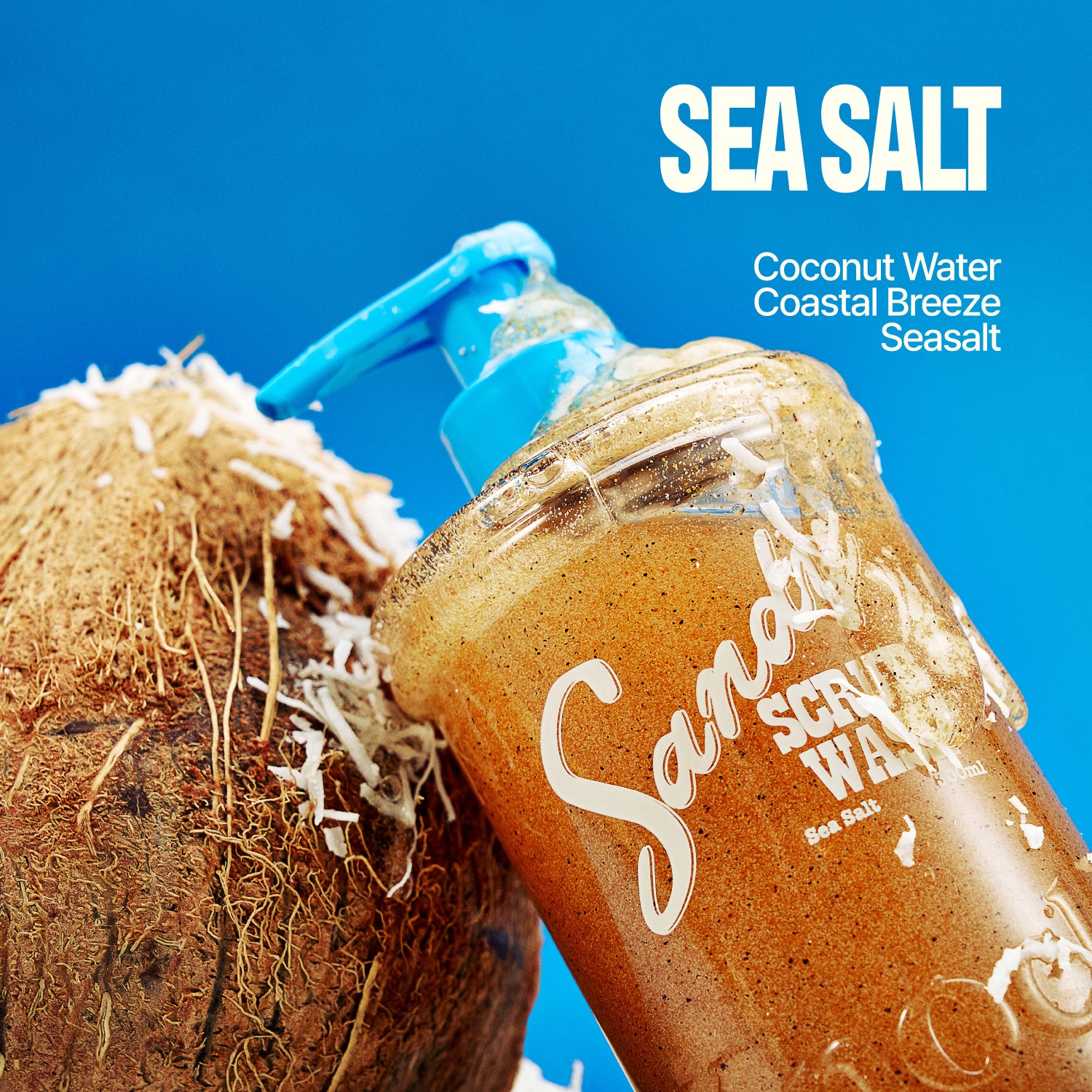 A bottle of SANDY SCRUB WASH with a blue pump sits next to a cracked coconut, coconut shavings scattered nearby. Text highlights SEA SALT, Coconut Water, Coastal Breeze, and Seasalts. Indulge in this invigorating body wash and coconut scrub blend for irresistibly smooth skin.