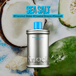 A silver bottle labeled MELT IN BODY TONER stands in front of coconut halves, a green fruit, and seashells. Text above reads SEA SALT with hashtags #CoconutWater #CoastalBreeze #Seasalt. Experience the refreshing feel of this moisturizing body toner that absorbs instantly for a long-lasting scent.