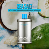 A silver bottle labeled MELT IN BODY TONER stands in front of coconut halves, a green fruit, and seashells. Text above reads SEA SALT with hashtags #CoconutWater #CoastalBreeze #Seasalt. Experience the refreshing feel of this moisturizing body toner that absorbs instantly for a long-lasting scent.