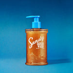 A bottle of SANDY SCRUB WASH, made with sea salt and coconut scrub, displays a blue pump top and orange-brown scrub inside, set against a blue background—ideal for achieving smooth skin.