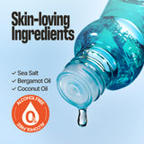 A close-up of a transparent bottle pouring clear blue liquid, with a single drop dripping below the opening. Large text reads 'Skin-loving Ingredients' with checked items for Sea Salt, Bergamot Oil, and Coconut Oil, plus an orange 'Alcohol Free' badge on a light blue background.