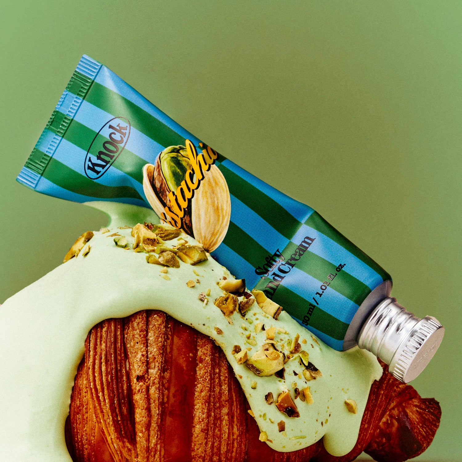 A striped tube labeled SOFTY HAND CREAM PISTACHIO, reminiscent of a serum-in-cream, rests on a croissant covered in green pistachio cream and chopped pistachios, against a green background.