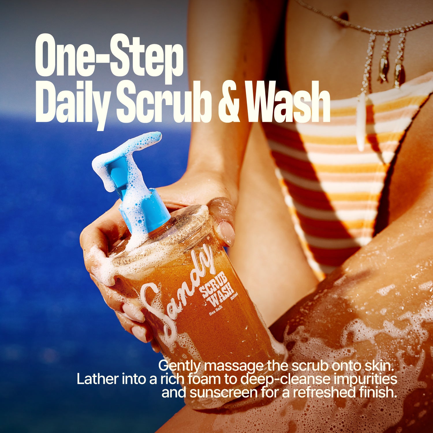 Hand holding a pump bottle labeled SANDY SCRUB WASH near a sun-dappled, sandy leg at the beach, with text promoting it as a one-step daily body wash and scrub for smoother skin.