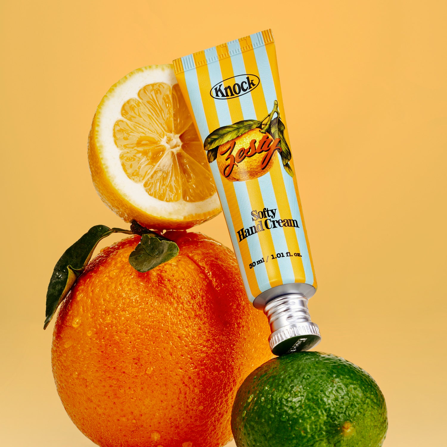 A tube of SOFTY HAND CREAM ZESTY, featuring a serum-in-cream formula for a silky finish, is balanced on a lime with an orange and half a lemon stacked behind it against a yellow background.