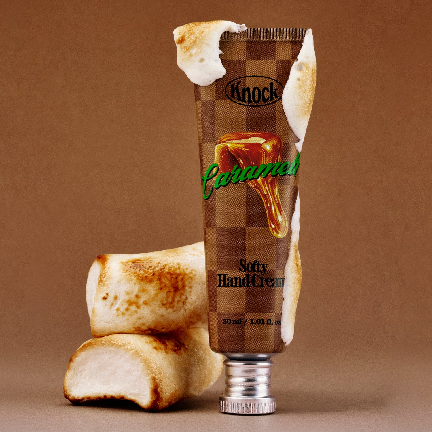 A tube labeled SOFTY HAND CREAM CARAMELT, adorned with a caramel and marshmallow design, is surrounded by toasted marshmallows, with melted cream dripping onto the tube. This decadent hand moisturizer delivers a silky finish, beautifully highlighted against a soft brown gradient background.