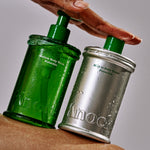 A hand rests on two bottles from the MELLOW BODY WASH & MELT IN TONER SET, one green and one silver, both adorned with water droplets and set against a neutral background.