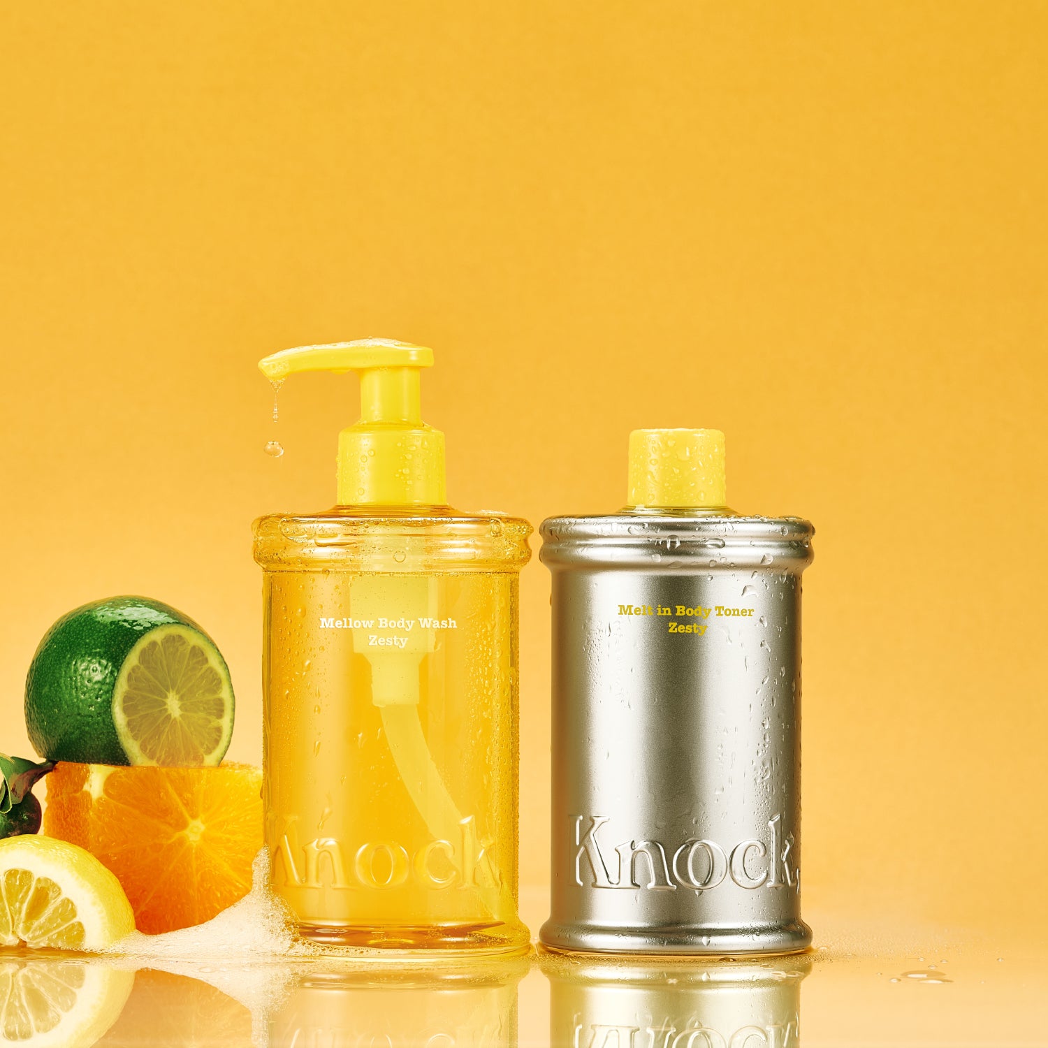 Two bottles, one with a pump and one metallic, on a yellow background with citrus fruits.